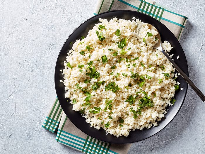 Best vegetables for weight loss - Cauliflower rice or couscous mixed with finely chopped parsley in a black bowl on a white concrete table, healthy eating, low calories food, horizontal view from above, flatlay, empty space